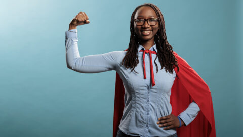 Jovem mulher negra, usa óculos e camisa azul e calça jeans. Faz posição de força e usa capa vermelha, simbolizando um herói.
