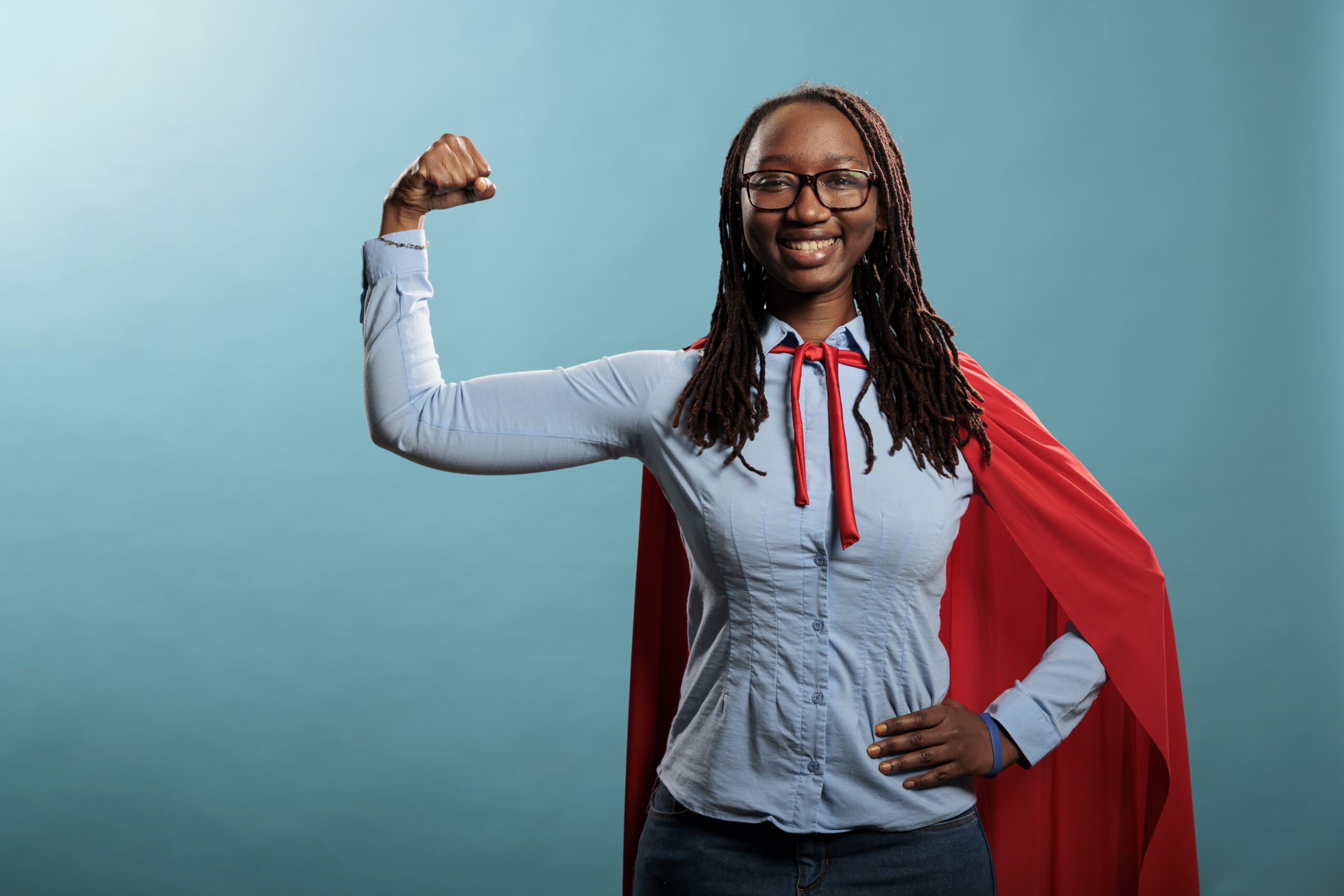 Jovem mulher negra, usa óculos e camisa azul e calça jeans. Faz posição de força e usa capa vermelha, simbolizando um herói.