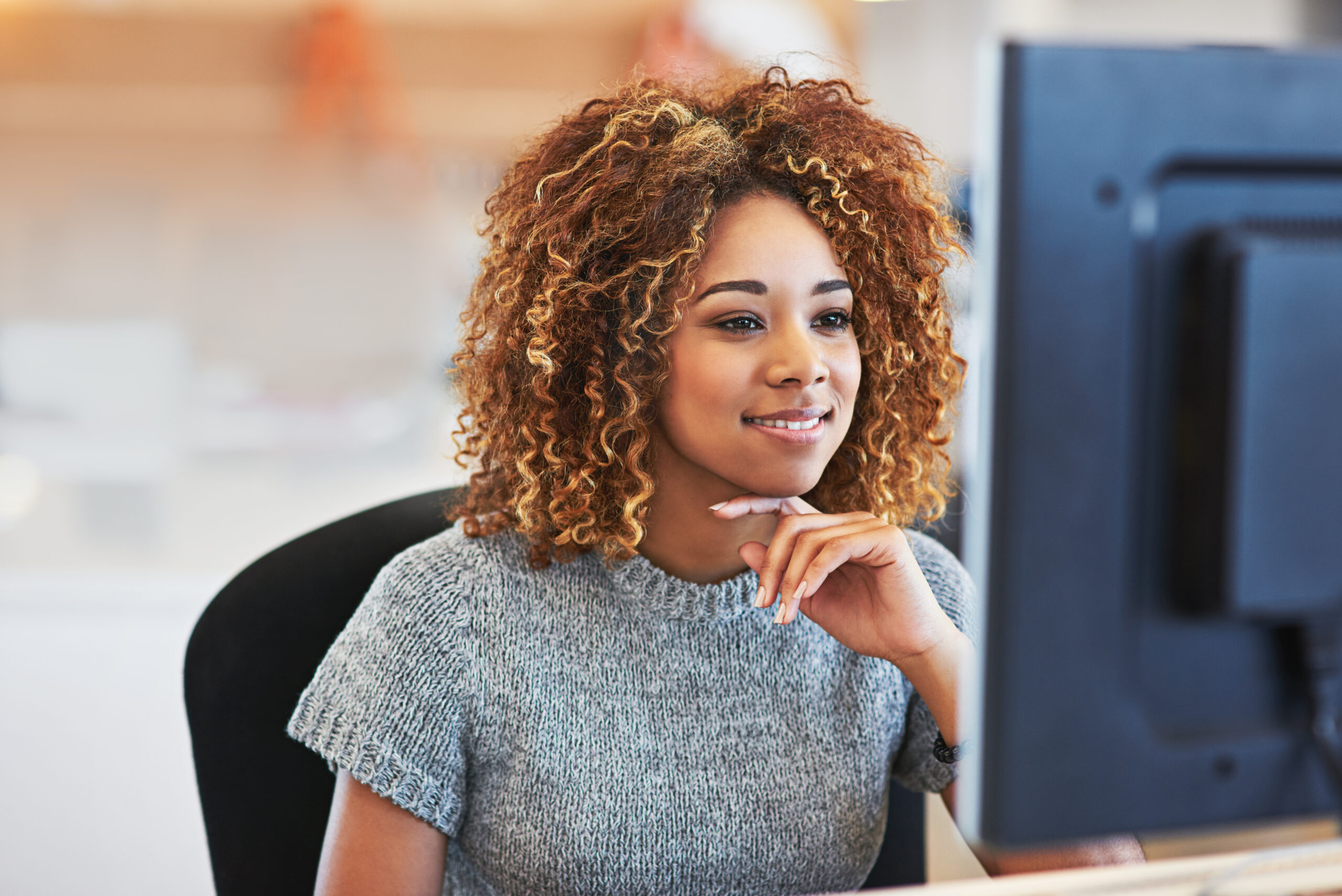 Jovem mulher negra, de cabelo médio e castanho claro. Usa blusa cinza e está sentada na cadeira e sorri enquanto olha o computador| Fonte da imagem: Envato